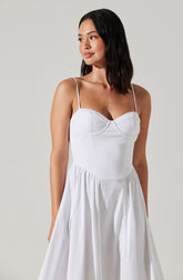 A young woman is wearing a white dress with thin spaghetti straps. The dress has a fitted bodice with a sweetheart neckline and subtle seam detailing. The skirt is full and gathered, creating a flowing, elegant silhouette.